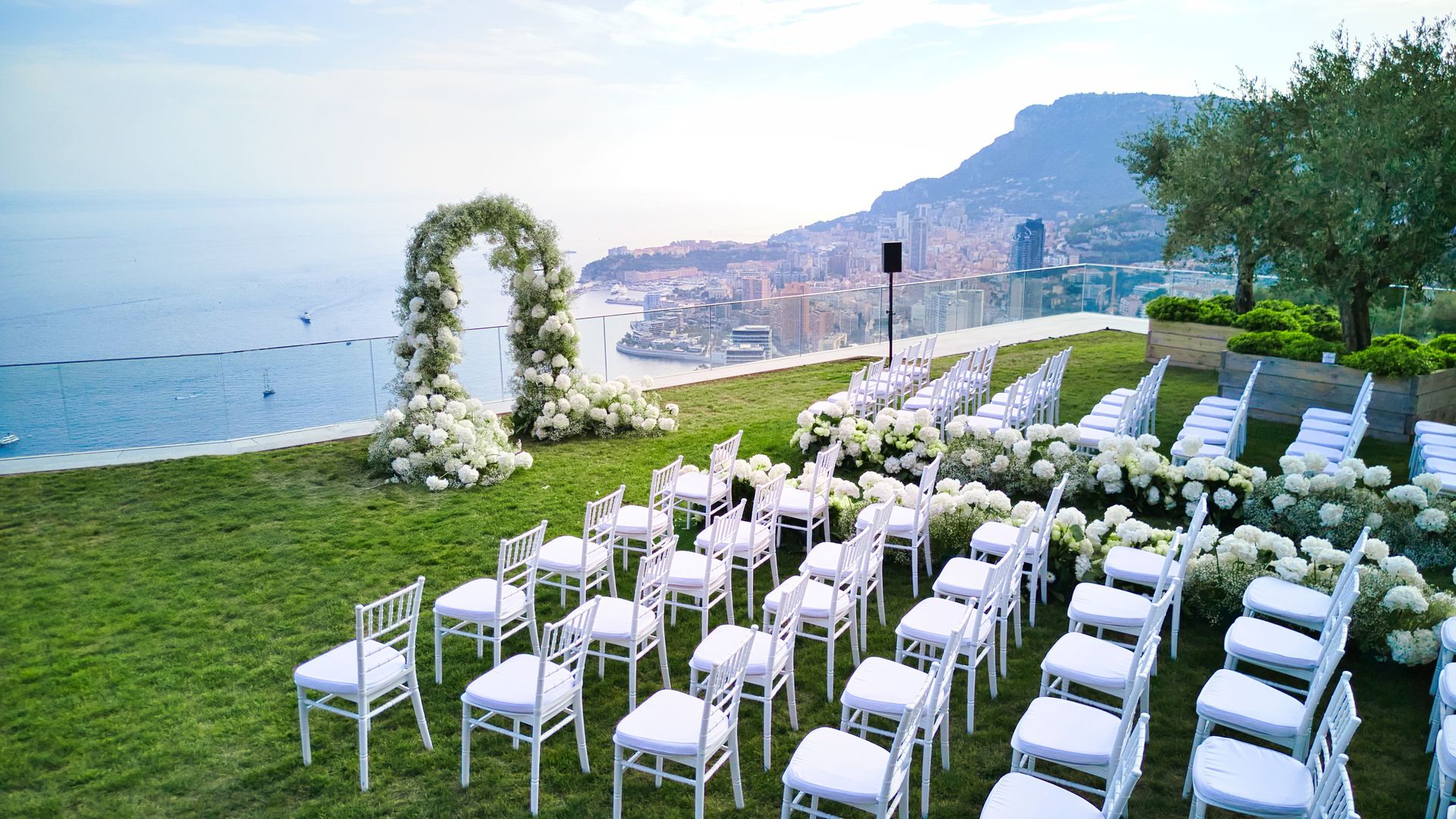 An idyllic cliffside ceremony with white floral arches and chairs, overlooking the breathtaking Mediterranean coastline and a picturesque cityscape below.