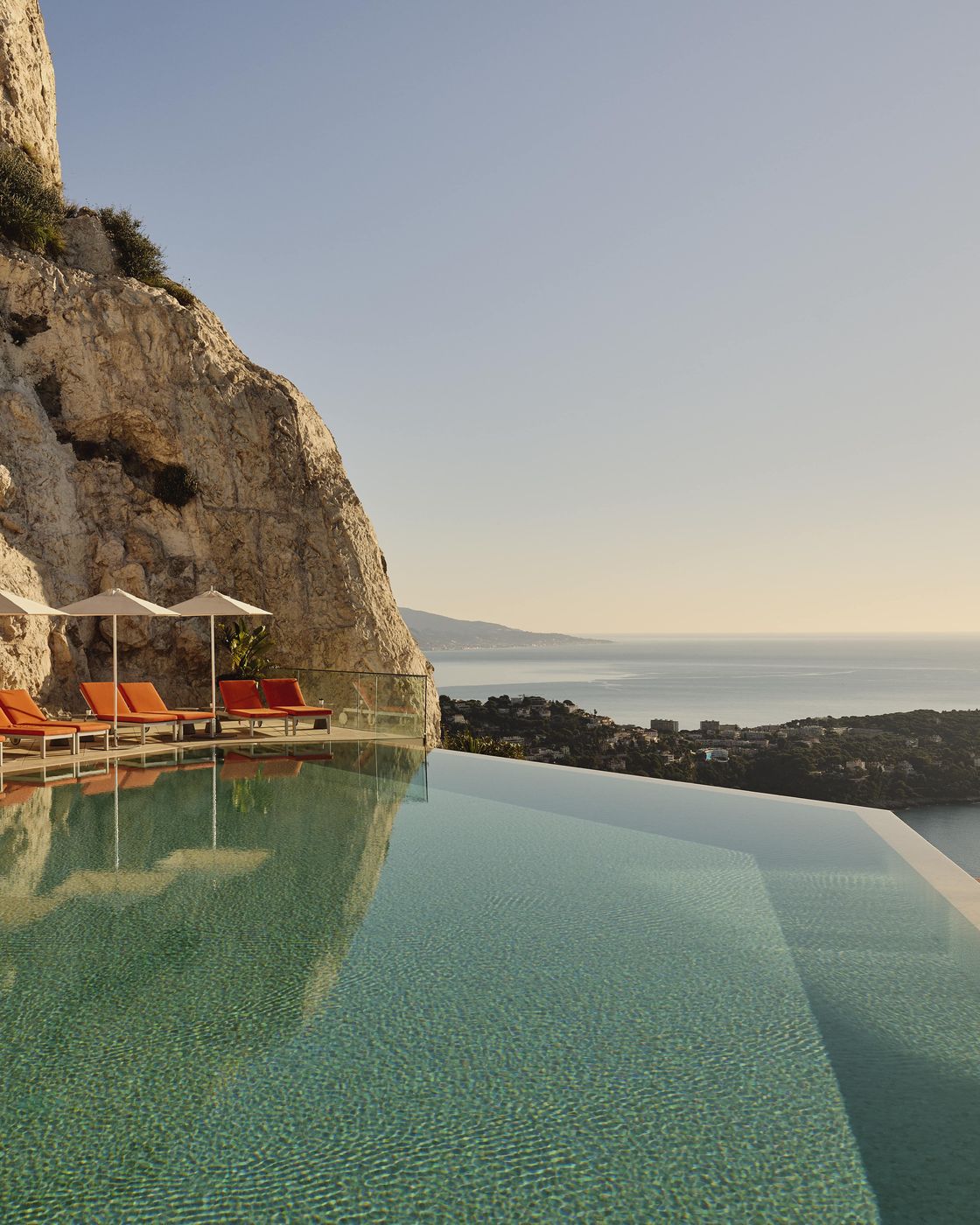 Infinity pool beside cliffside with orange loungers and umbrellas, overlooking coastline and sea under soft golden sunlight.