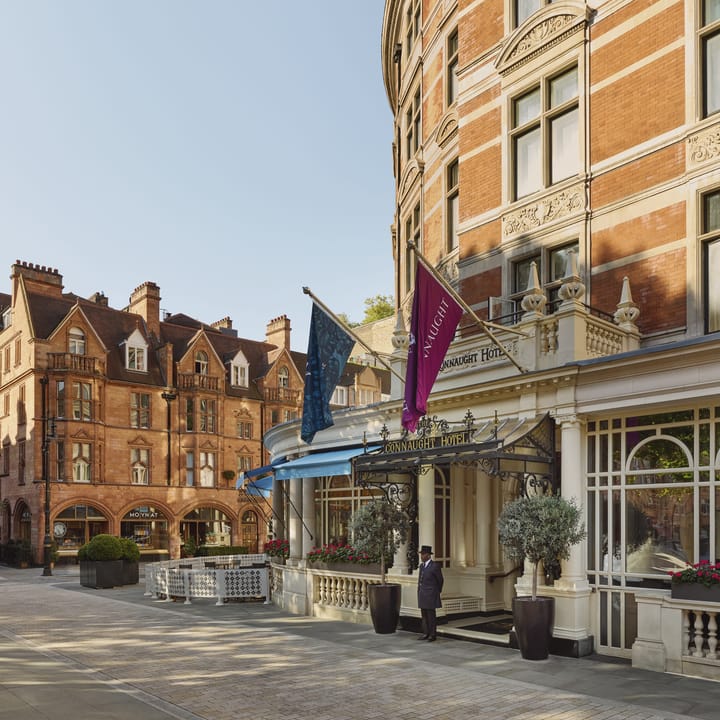 Elegant red-brick facade of The Connaught Hotel with flag-topped entrance, classic detailing, and doorman welcoming guests.