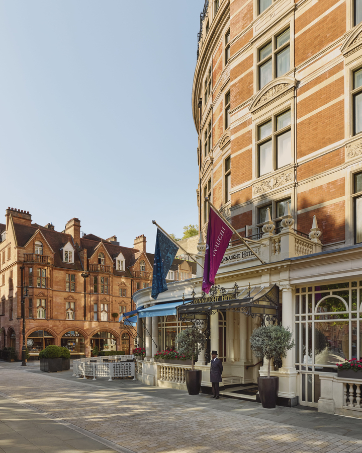 Elegant red-brick facade of The Connaught Hotel with flag-topped entrance, classic detailing, and doorman welcoming guests.