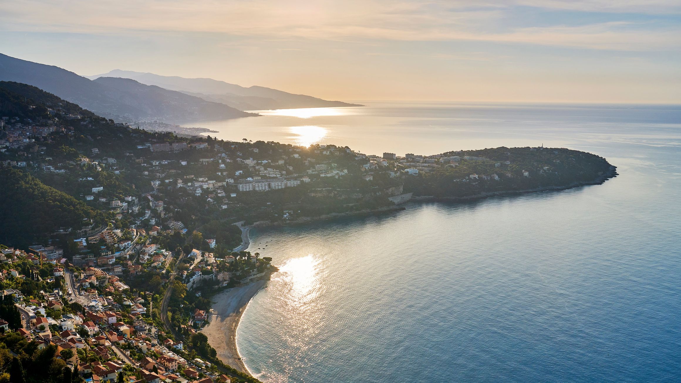 Sweeping coastal view of Cap Martin and the French Riviera at sunset, with shimmering sea, hillside villas, and soft golden light on the horizon.