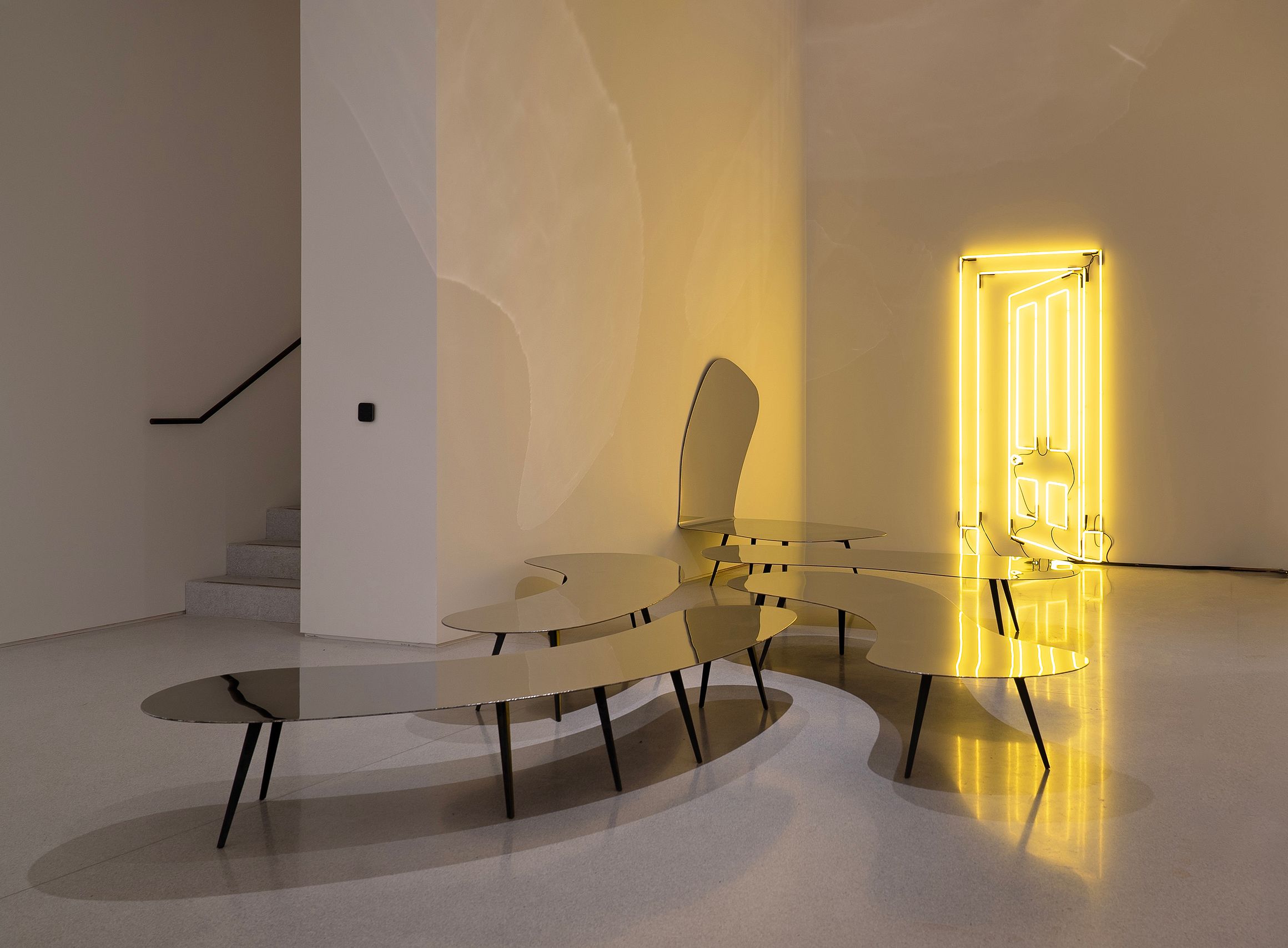 Contemporary interior with reflective sculptural tables, minimalist stairs, and a striking neon door art installation in warm yellow light.