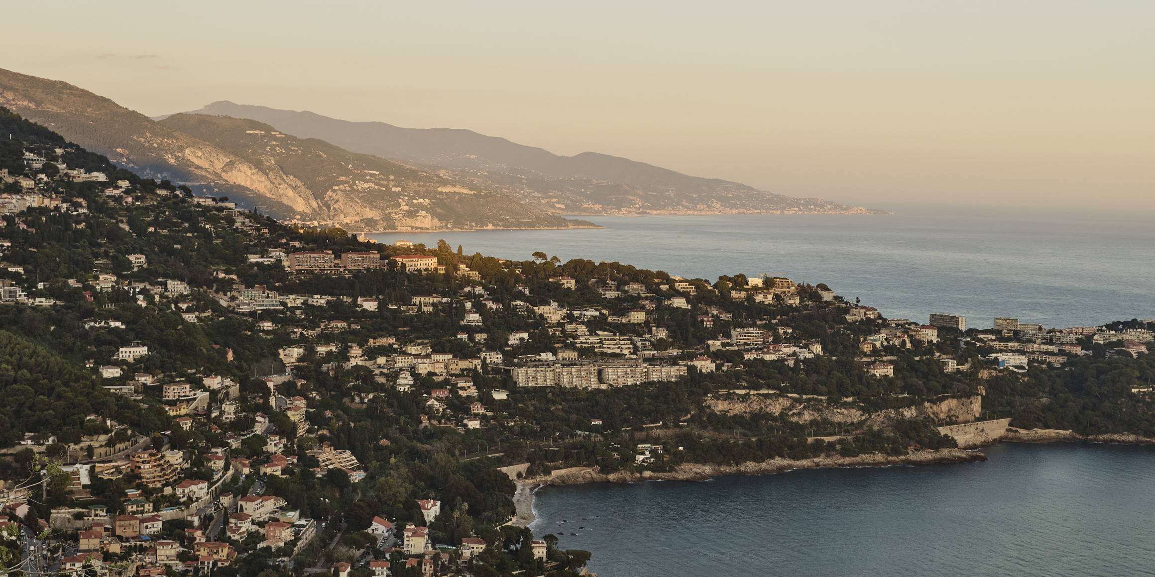 Aerial view of the French Riviera coastline at sunset, with hillside villas, lush greenery, and calm Mediterranean waters.