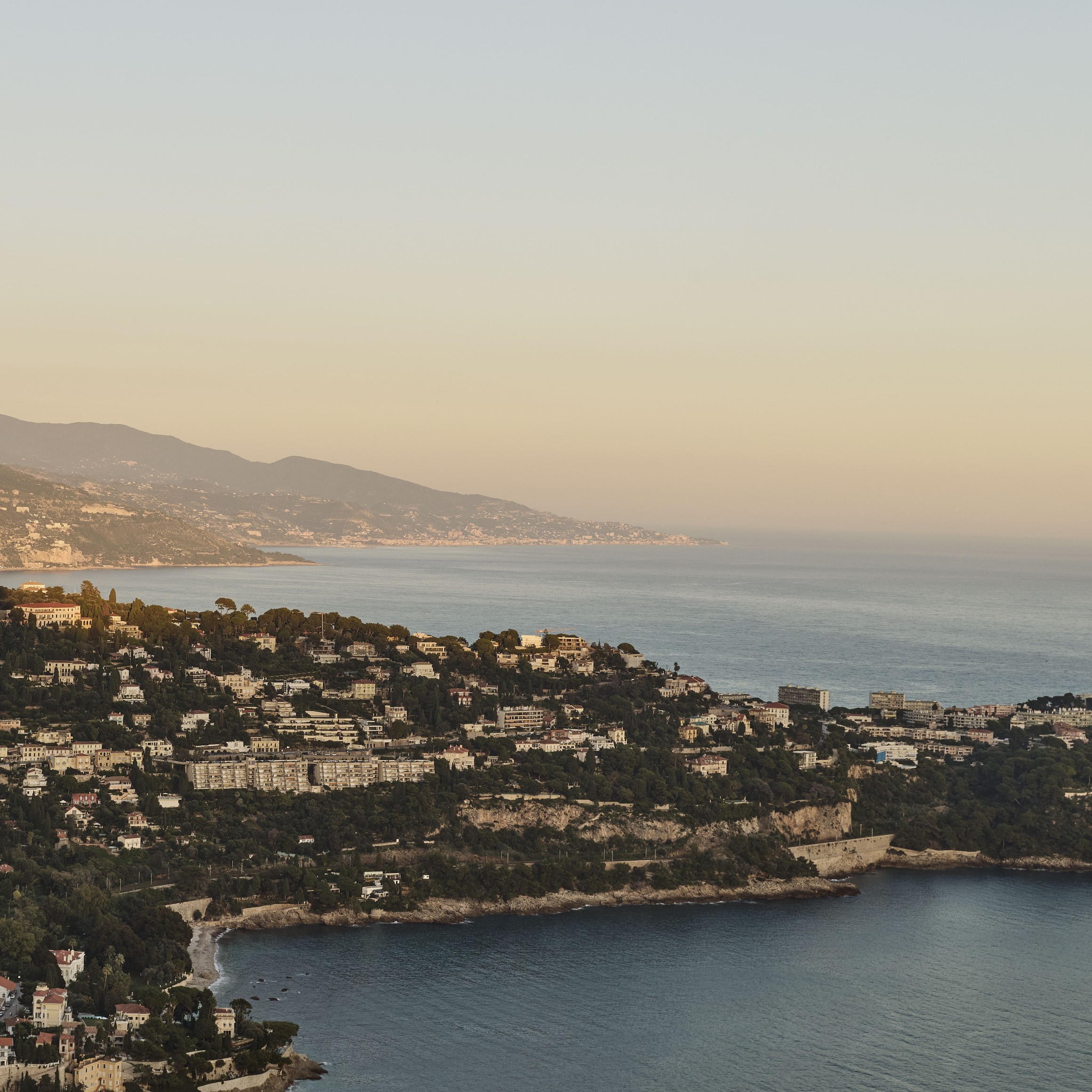 Aerial view of the French Riviera coastline at sunset, with hillside villas, lush greenery, and calm Mediterranean waters.